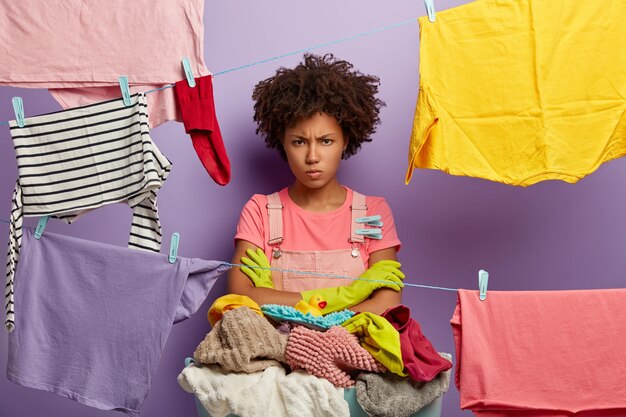 Cross unhappy housewife stands with arms folded, hands washed laundry, angry with much duties about house, uses clothespegs, isolated over violet wall. People, housework and washing concept.