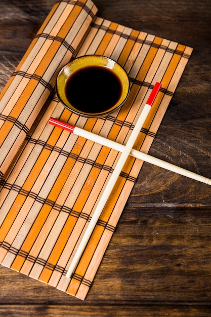 Free photo crossed chopsticks and bowl of soya sauces on placemat over the wooden desk