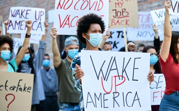 도시 거리에서 시위하는 동안 보호용 안면 마스크를 쓴 사람들의 군중 초점은 '미국을 깨워라'라는 문구가 적힌 현수막을 들고 있는 흑인 여성에 대한 것입니다.