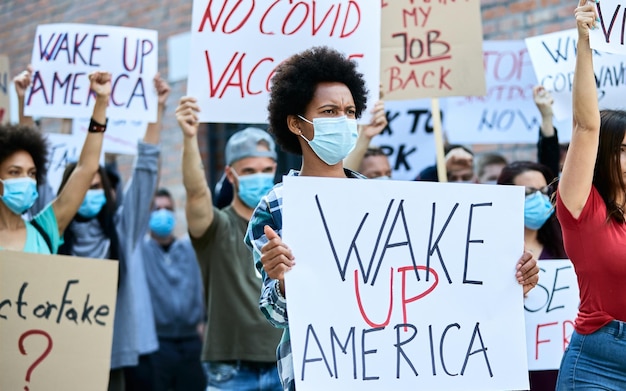 Толпа людей в защитных масках во время акции протеста на улицах города. В центре внимания чернокожая женщина, держащая транспарант с надписью «Проснись, Америка».