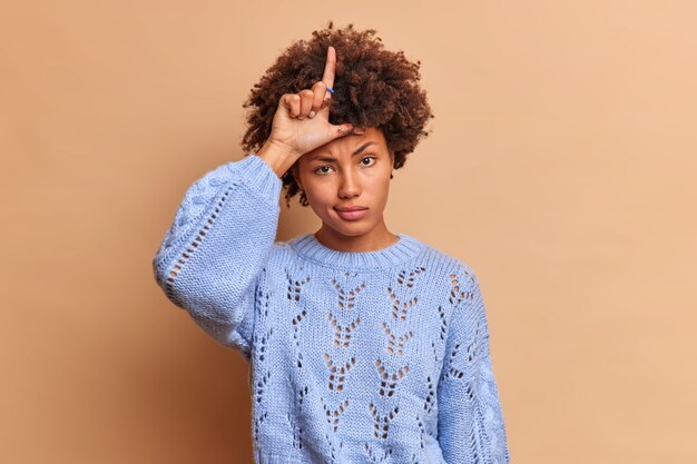 Cruel rude serious woman makes lose gesture keeps finger raised over forehead insults someone makes fun of people wears blue sweater isolated over beige wall