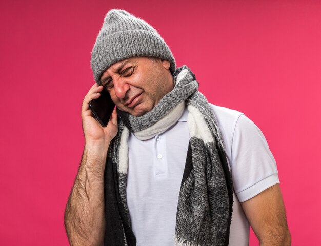 crying adult ill caucasian man with scarf around neck wearing winter hat talking on phone isolated on pink wall with copy space