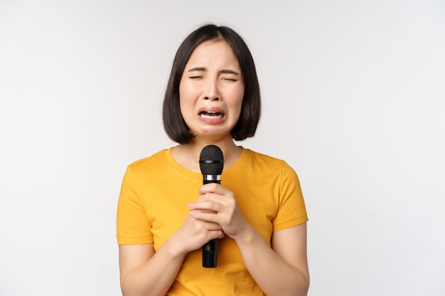 Crying asian girl singing heartbroken in microphone holding mic and grimacing upset standing over white background Copy space