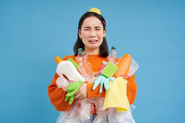 더러운 라텍스 장갑을 사용하는 빈 플라스틱 병을 들고 울고 있는 아시아 여성은 재활용 쓰레기에 대해 울고 있습니다.