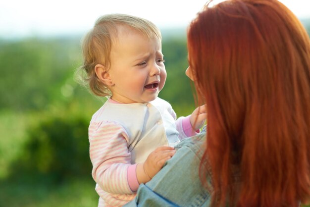 Crying daughter sitting on mother's hands during sunny day in the field Young mom trying to calm down little baby talking to her Woman having red hair wearing jeans shirt