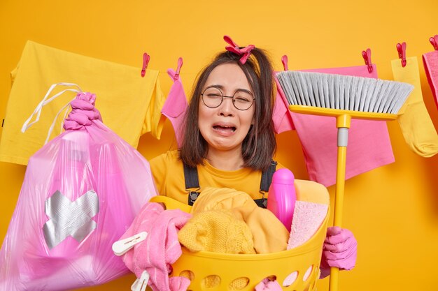 울고 좌절 한 아시아 여성이 바닥 청소를 위해 세제 빗자루 병이 든 폴리에틸렌 가방을 들고 낙담 한 표정을 지으며 둥근 안경을 쓰고 고무 장갑을 끼고 집에서 세탁을합니다.