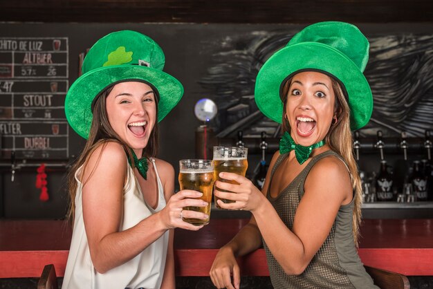 Crying happy women in Saint Patricks hats clanging glasses of drink at bar counter