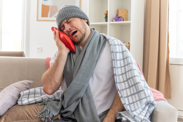 Crying ill slavic man with scarf around neck wearing winter hat wrapped in plaid holding and looking at hot water bottle sitting on couch at living room