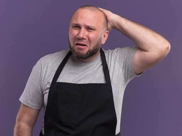 Crying middle-aged male barber in uniform putting hand on behind head isolated on purple wall