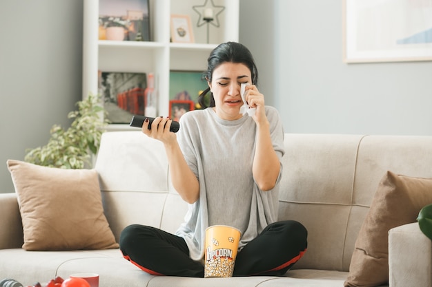 Crying wiping eye with napkin young girl holding tv remote sitting on sofa behind coffee table in living room