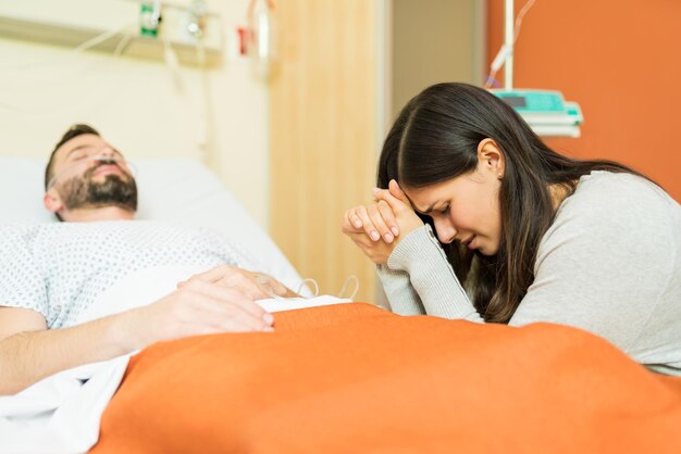 Плачущая женщина молится за пациента мужского пола, сидя у кровати в больнице во время визита
