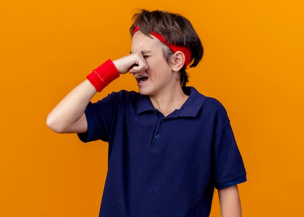 Crying young handsome sporty boy wearing headband and wristbands with dental braces wiping tears isolated on orange wall