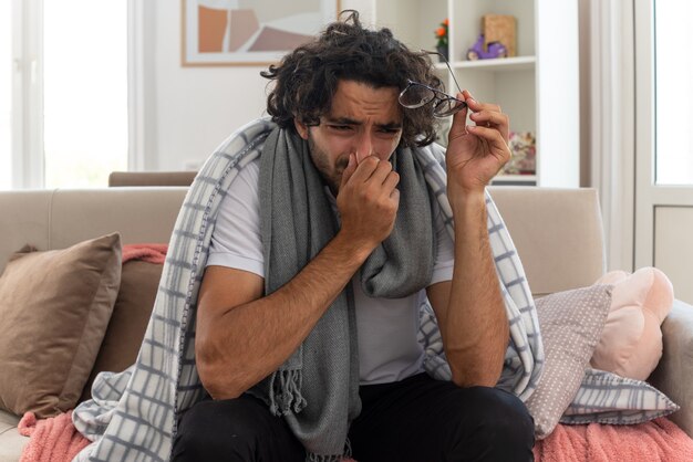 crying young ill caucasian man wrapped in plaid with scarf around his neck holding optical glasses and putting hand on nose sitting on couch at living room