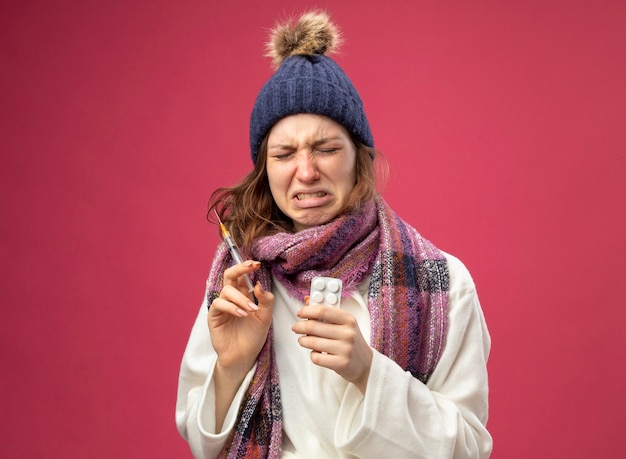 Crying young ill girl wearing white robe and winter hat with scarf holding syringe with pills isolated on pink