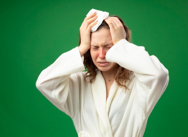 Crying young ill girl with closed eyes wearing white robe grabbed head