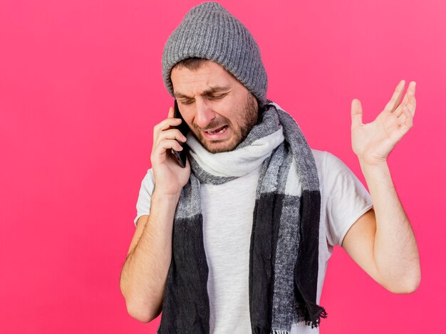 Crying young ill man wearing winter hat with scarf speaks on phone and raising hand isolated on pink background