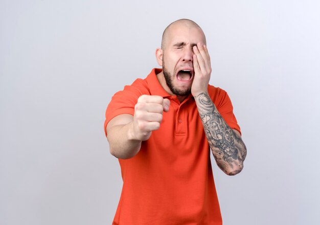 crying young sporty man holding out fist at camera and covered eye isolated on white