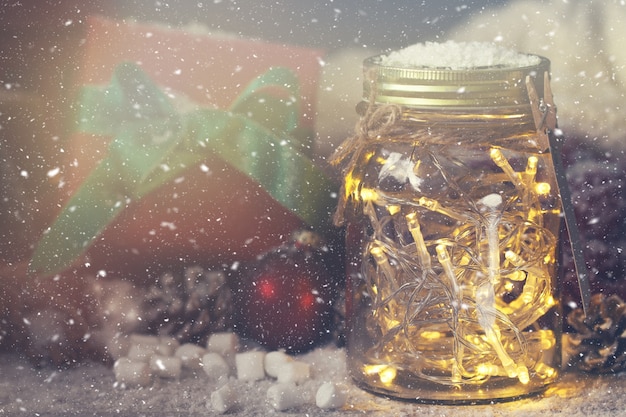 Кристалл банку с огнями с подарком рядом с ним в то время как снег идет
