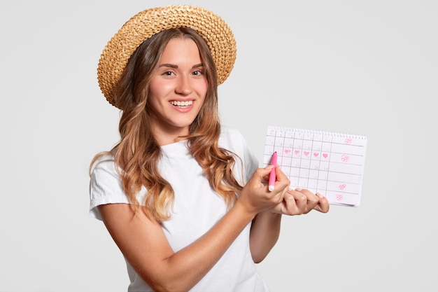 Cuacasian woman has charming smile, holds periods calendar, marks with marker day of starting menstruation