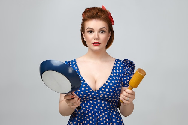 Culinary and cooking. Amazed beautiful young Caucasian housewife wearing vintage dotted dress with low cut staring at camera with mouth opened, holding frying pan and rolling pin