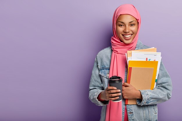 Culture, religion and studying concept. Glad Muslim woman with toothy smile, carries notebook with papers, takeaway coffee