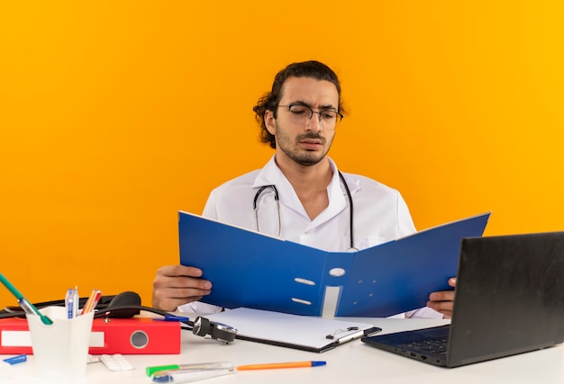 Молодой мужчина-врач в медицинских очках в медицинском халате со стетоскопом сидит за столом