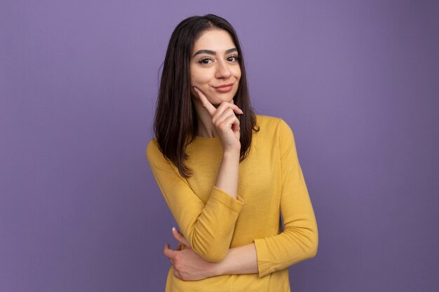 Cunning young pretty caucasian girl putting hand on chin isolated on purple wall with copy space