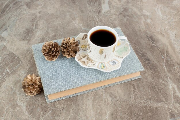 Cup of aromatic coffee on top of book with pinecones