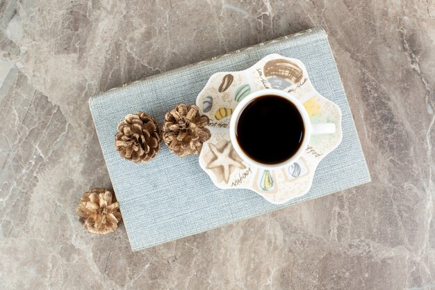 Cup of aromatic coffee on top of book with pinecones