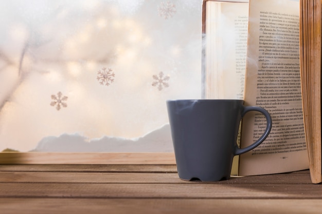 Cup and book on wood table near bank of snow and snowflakes 