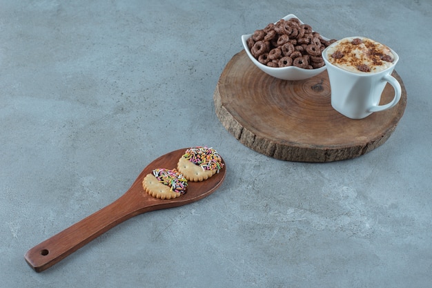 A cup of cappuccino and a bowl of corn ring on a board next to spoon, on the blue background.