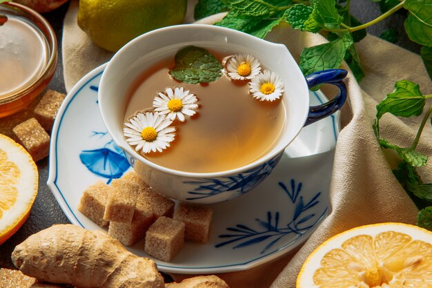 Cup of chamomile tea with slices of lemon, ginger, brown sugar cubes and green leaves in a saucer on grey and picnic cloth background, close-up.