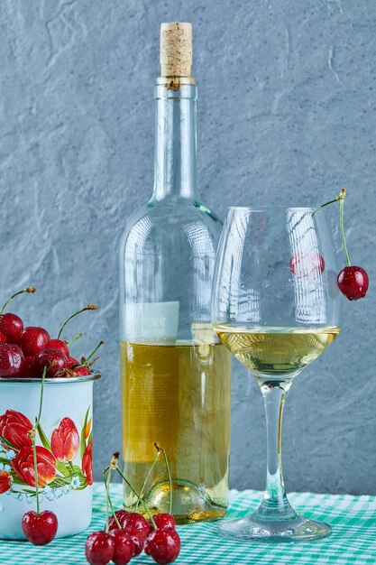 Cup of cherries, bottle of white wine and glass on blue surface