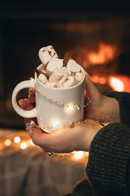 A cup of cocoa with marshmallows in female hands by the fireplace