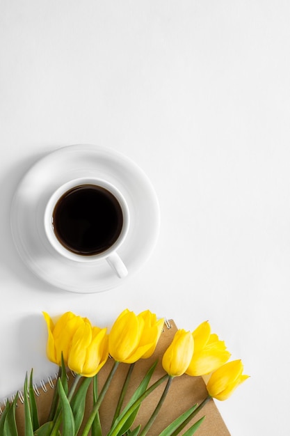 Free photo a cup of coffee bouquet of yellow tulips and a notebook on a white background