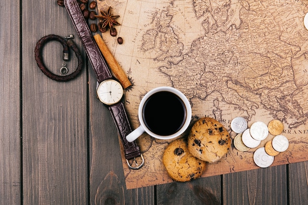 Cup of coffee, old yellow map, glasses, coins, leather case, camera, watch, compasses, coffee beans, other spices and cookies lie on wooden floor