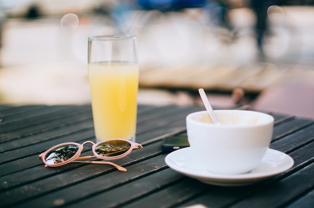 Foto gratuita tazza di caffè su un piattino con un succo d'arancia e un paio di occhiali da sole su un tavolo di legno