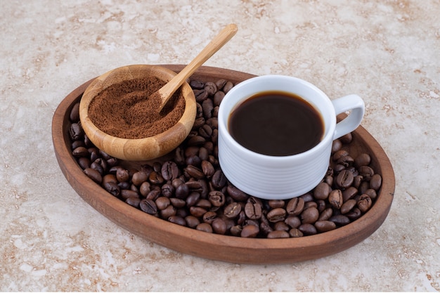 A cup of coffee and a small bowl of ground coffee powder on a pile of coffee beans in a tray 