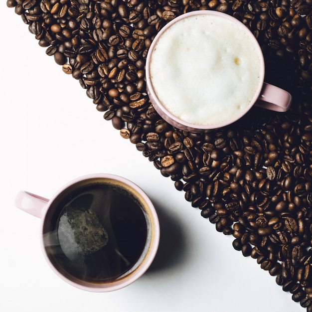 Tazza di caffè si leva in piedi sul tavolo bianco e tazza di latte sul tavolo coperto di grani di caffè