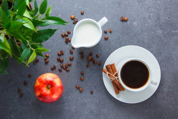 A cup of coffee with apple, dry cinnamon, plant, milk on gray surface, top view.