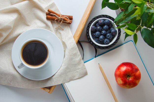 A cup of coffee with blueberries, apple, dry cinnamon, plant, pencil and notebook top view on a white surface