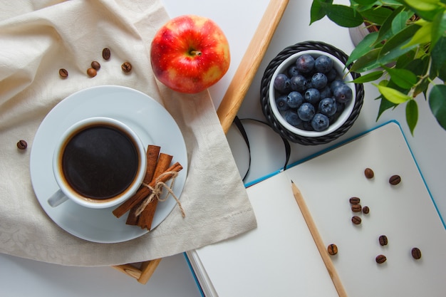 A cup of coffee with blueberries, apple, dry cinnamon, plant, pencil and notebook top view on a white surface