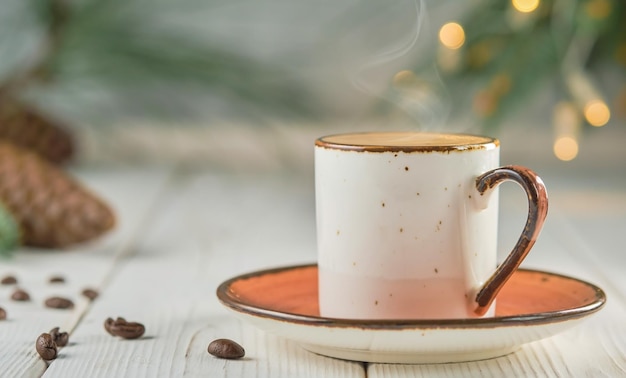 Cup of coffee with natural steam on a white wooden table, selective focus. Christmas background with copy space. Hot coffee drink concept. Christmas tree with a garland on the table. New year concept