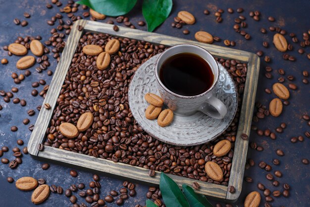 A cup of coffee with roasted coffee beans and coffee bean shaped cookies on dark surface