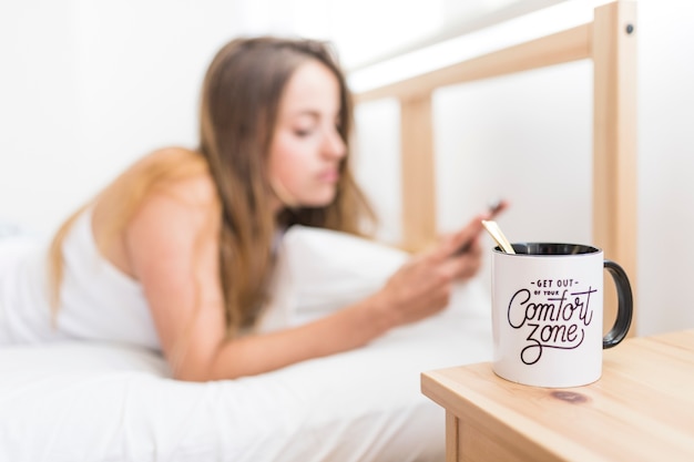 Чашка кофе на деревянном столе перед женщиной, лежащей на кровати с помощью мобильного телефона