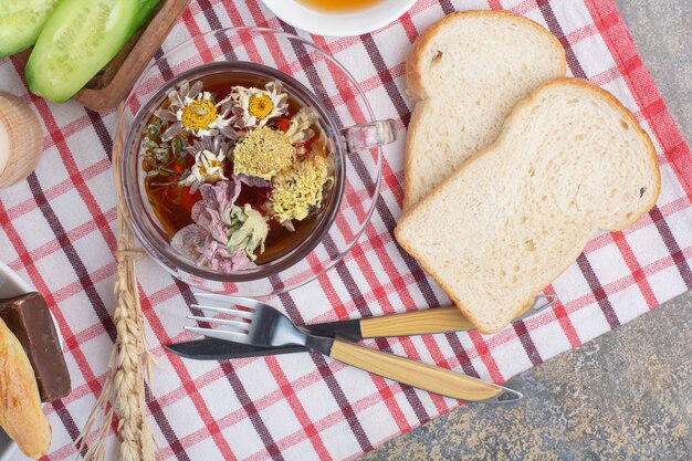 Cup of herbal tea, bread slices and cutlery on tablecloth.