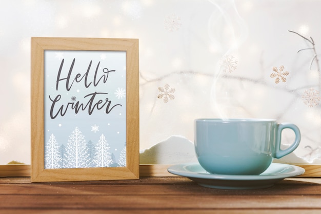 Free photo cup near frame with hello winter title on wood table near bank of snow
