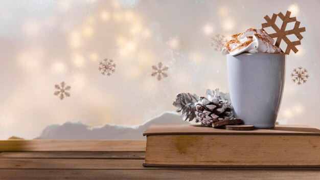 Cup, snag and book on wood table near bank of snow, snowflakes and fairy lights 