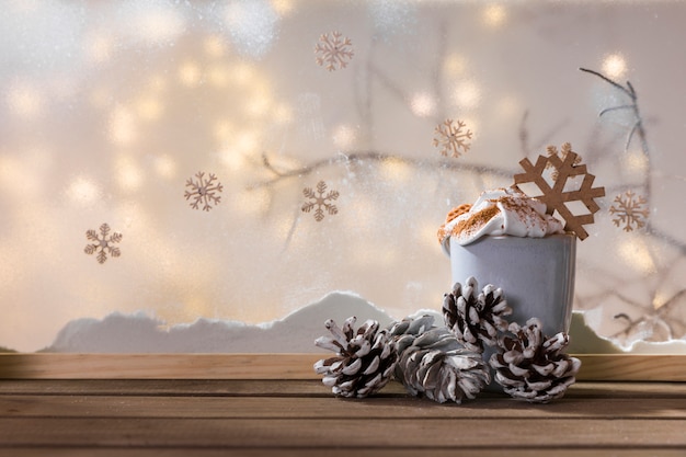 Cup and snags on wood table near bank of snow, plant twig, snowflakes and fairy lights