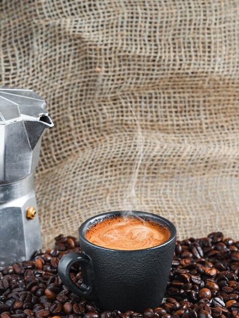 コーヒーポットの隣にある、焙煎したコーヒー豆の中に泡が入った強い芳香のエスプレッソコーヒーのカップ